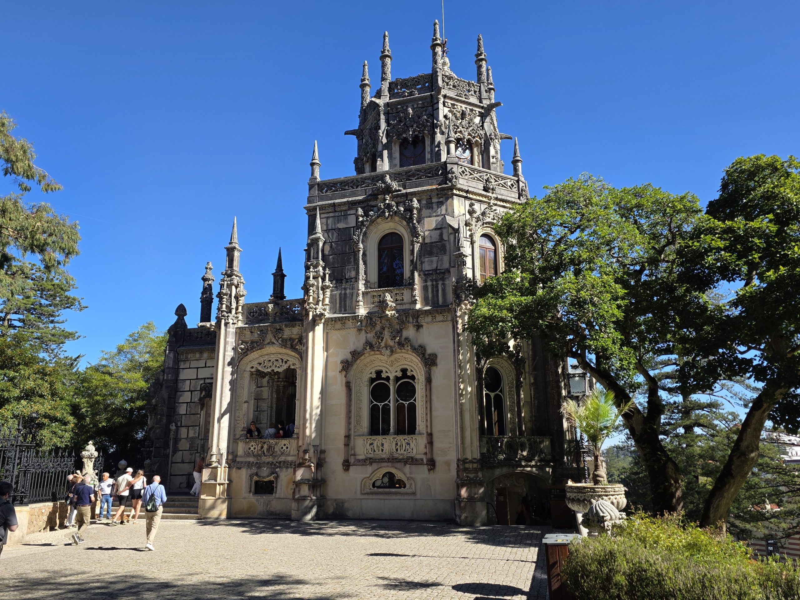 Side of Regaleira Palace with an ornate tower, carved windows, and a mature tree.