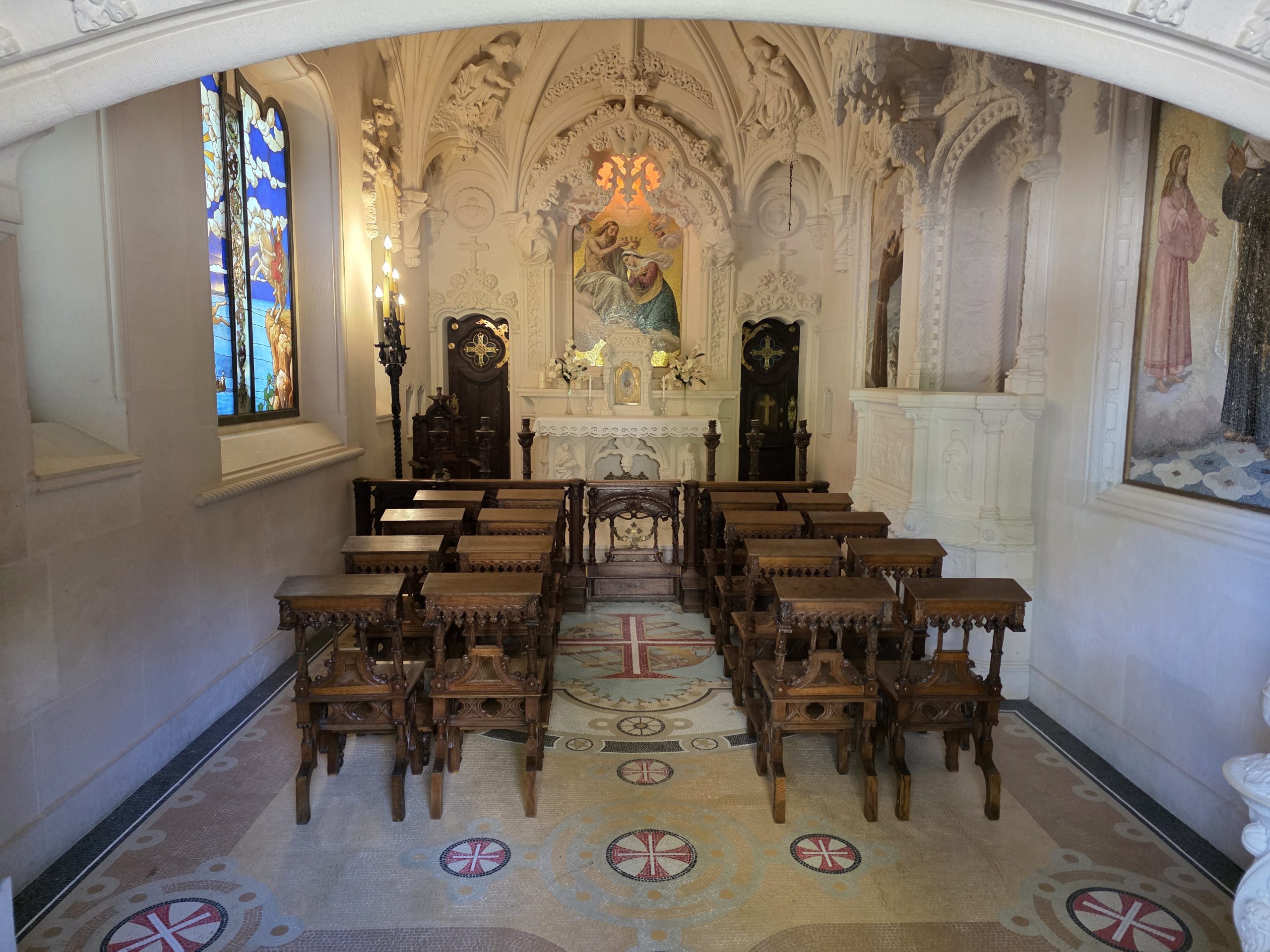 Chapel interior with carved wooden seats, patterned flooring, stained glass, and an ornate altar.