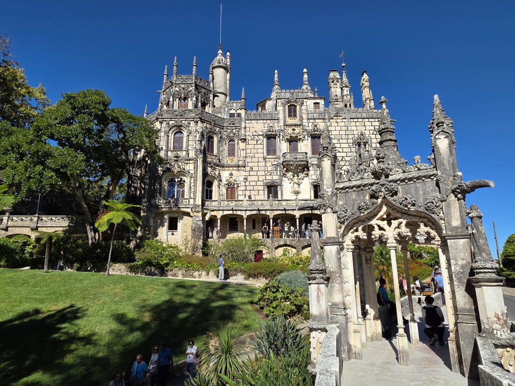 Ornately carved Regaleira Palace overlooking a green lawn with an elaborate stone gateway in the foreground.