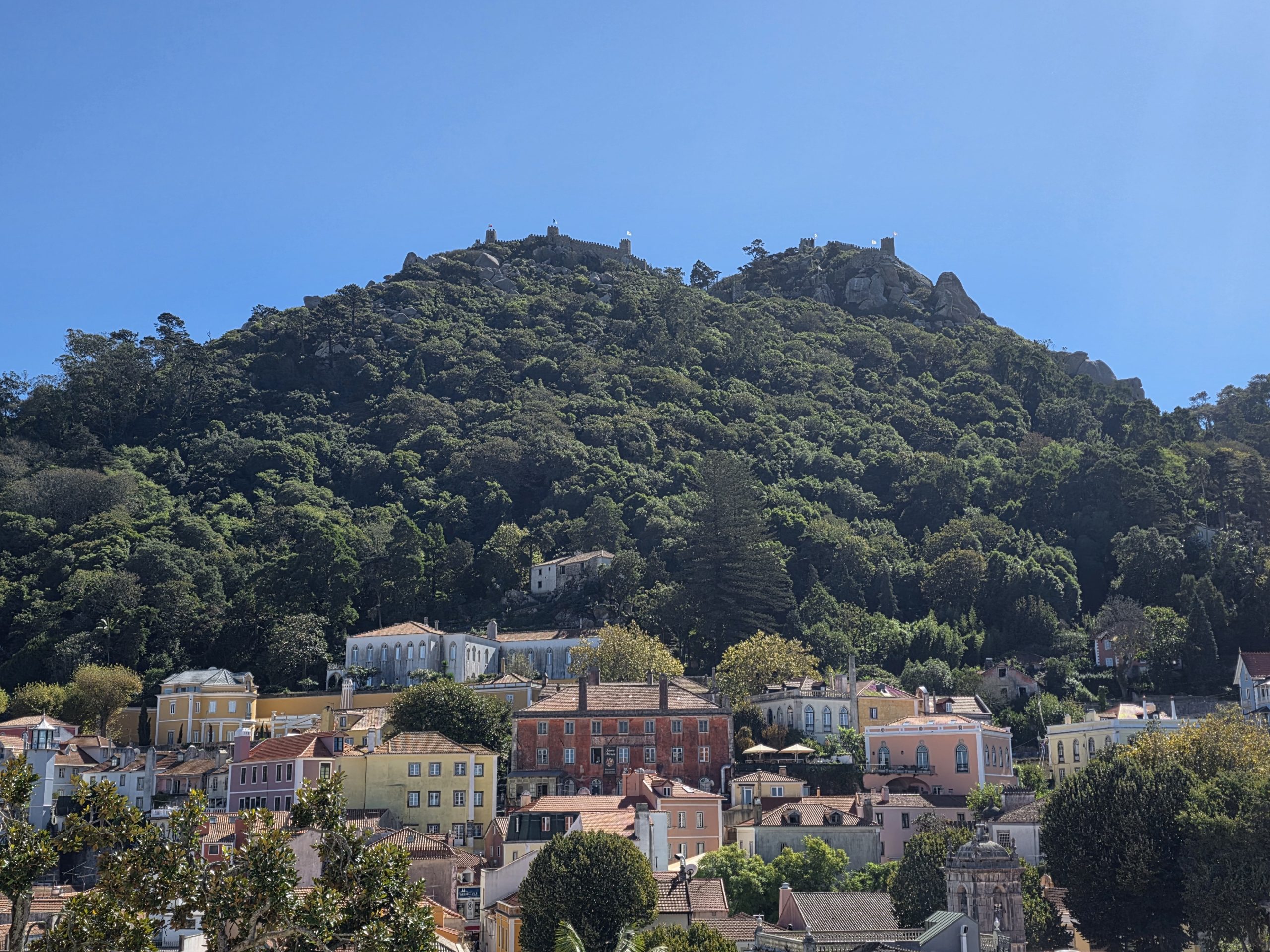 Colourful buildings and tiled roofs below a wooded hillside topped by castle walls in Sintra.